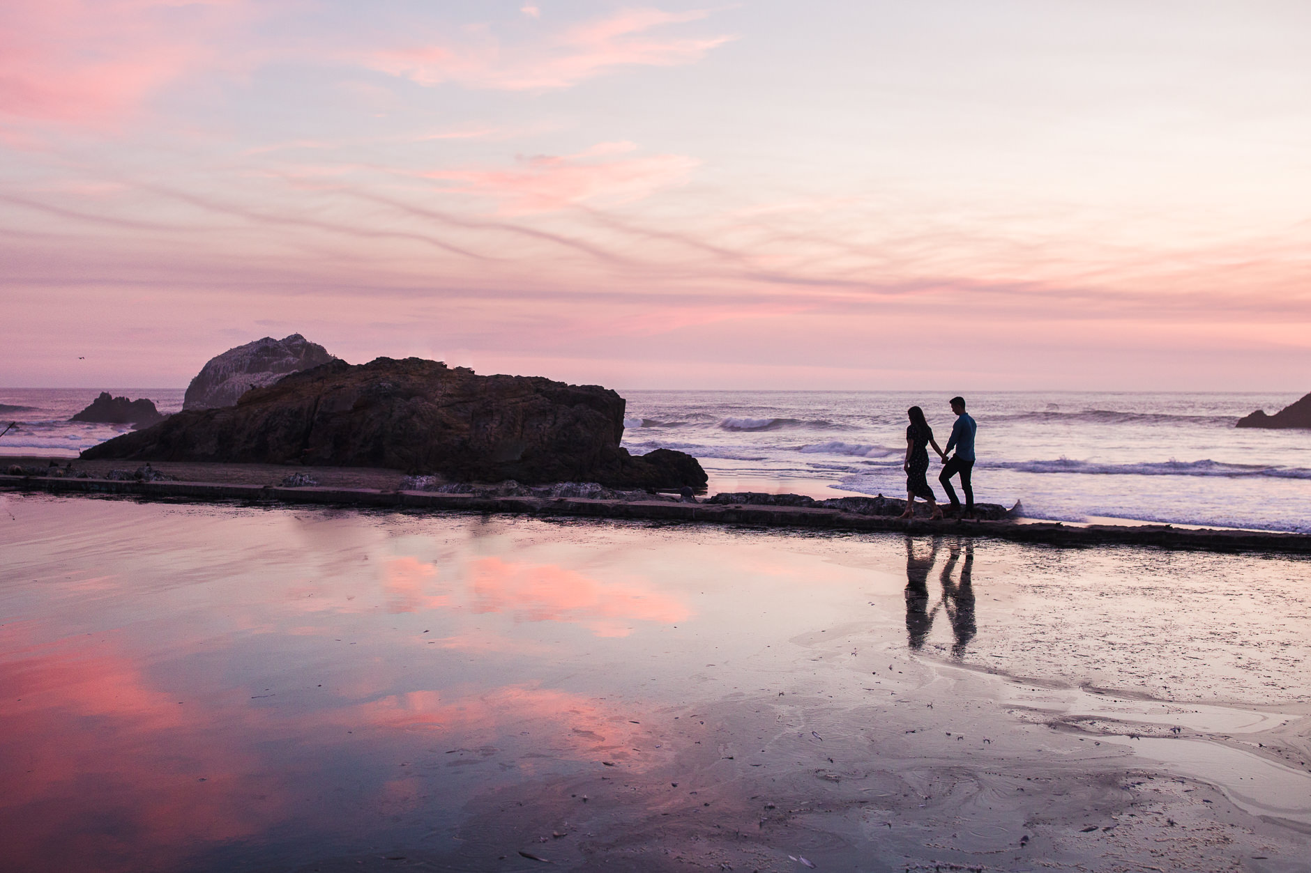 Silhouetted couple walking along ruins of Sutro Bath at sunset with reflection