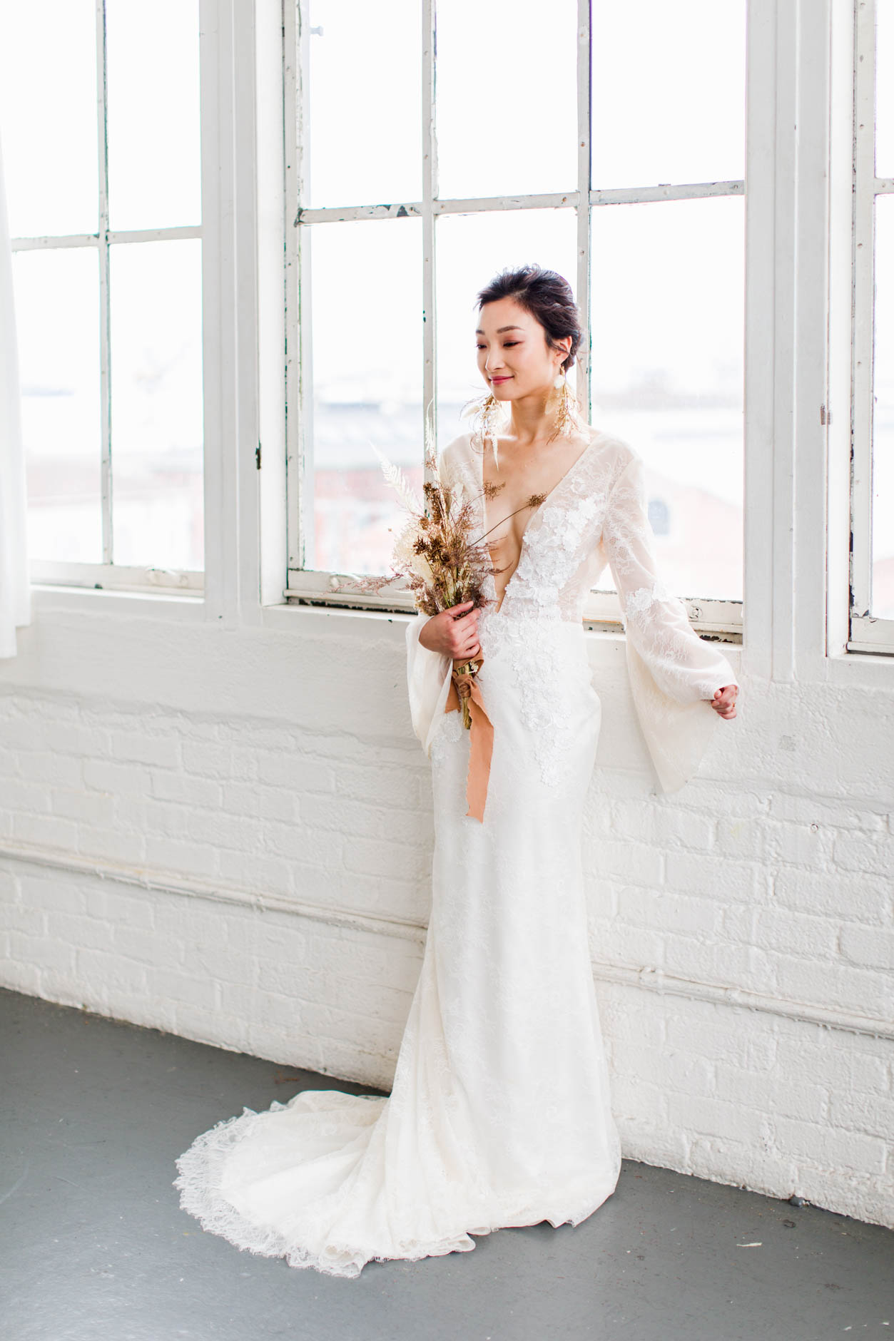 Bride with dried floral bouquet and lace bell sleeves standing in front of windows in modern warehouse wedding inspiration
