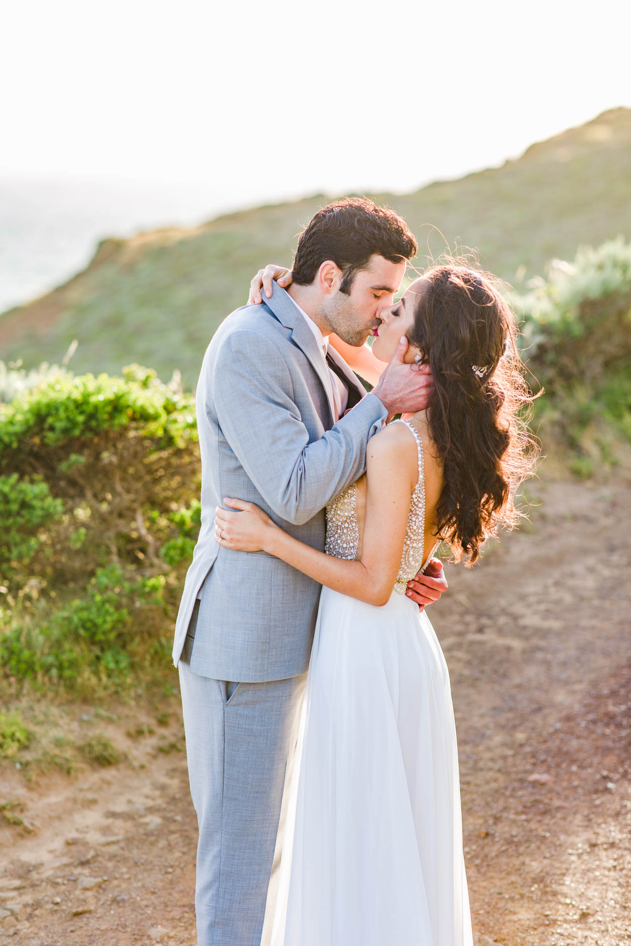 Bride wearing white sequined dress and groom wearing grey suit kissing in Marin Headlands