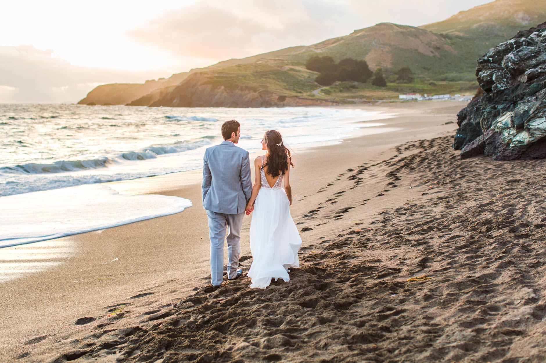 Bride and groom walking away from camera on Rodeo Beach at sunset