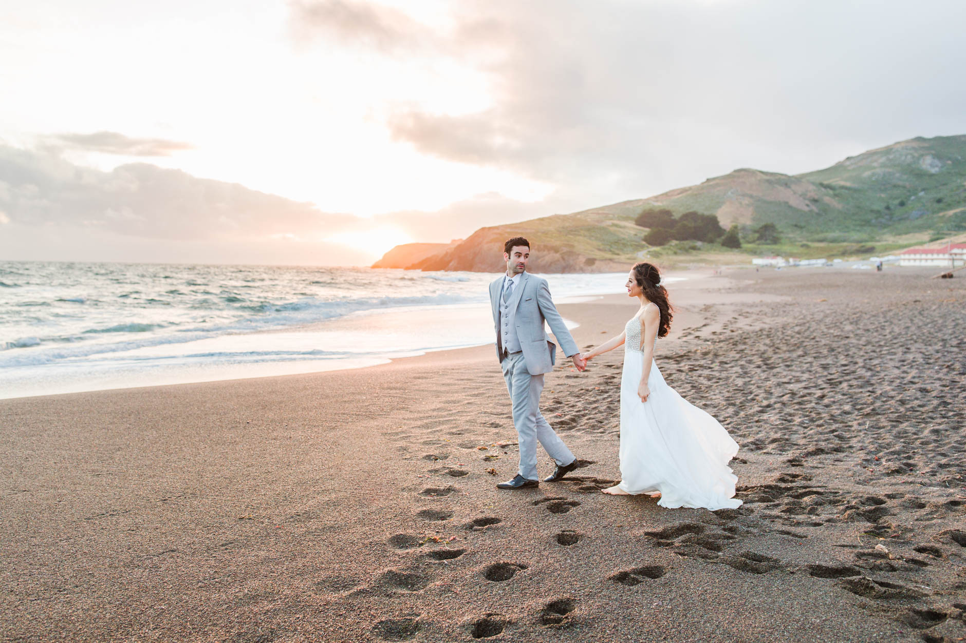 Groom looking back at bride while walking on Rodeo Beach at sunset